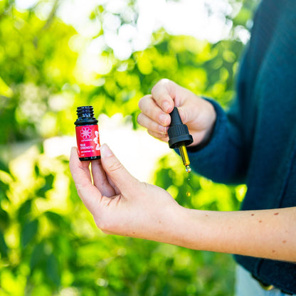 A person holds a True Strength Anointing Oil bottle and dropper, sunlit green leaves blurred behind